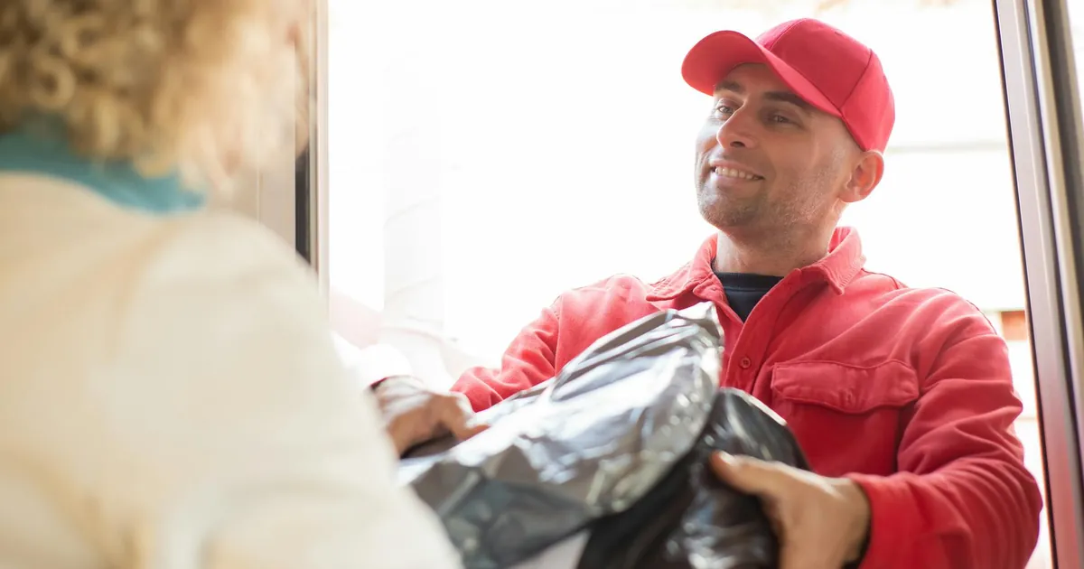 A smiling delivery man in red uniform hands over a package to a customer by the door.