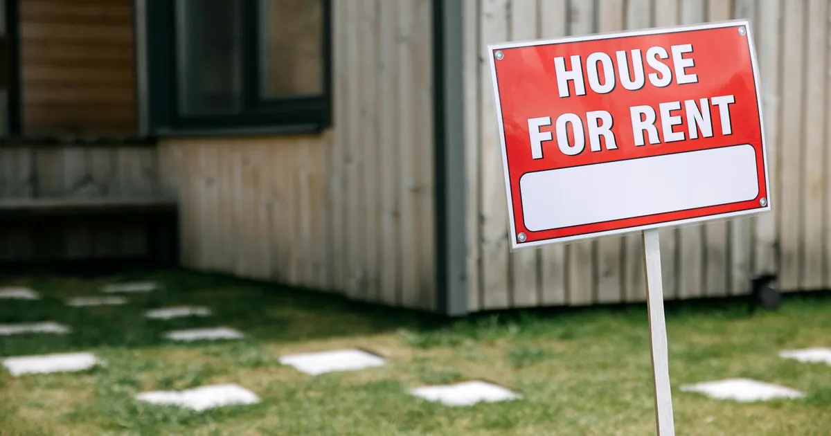 A modern wooden house with a prominent 'House for Rent' sign in the green yard.