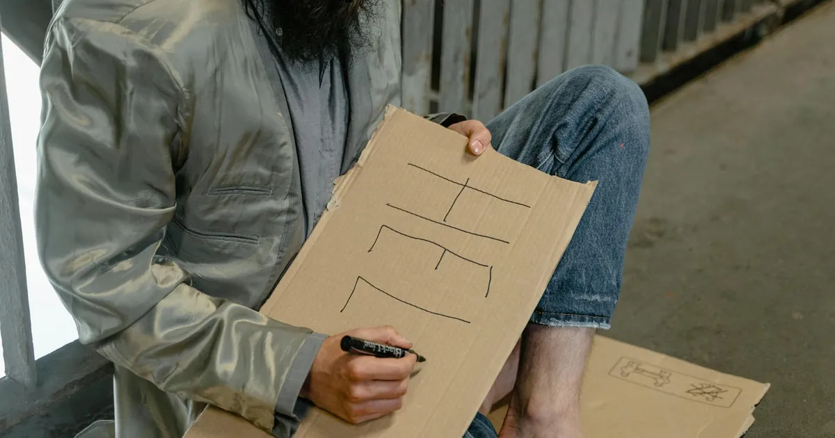 A homeless man writing 'Help' on a cardboard sign in an urban environment, reflecting poverty.