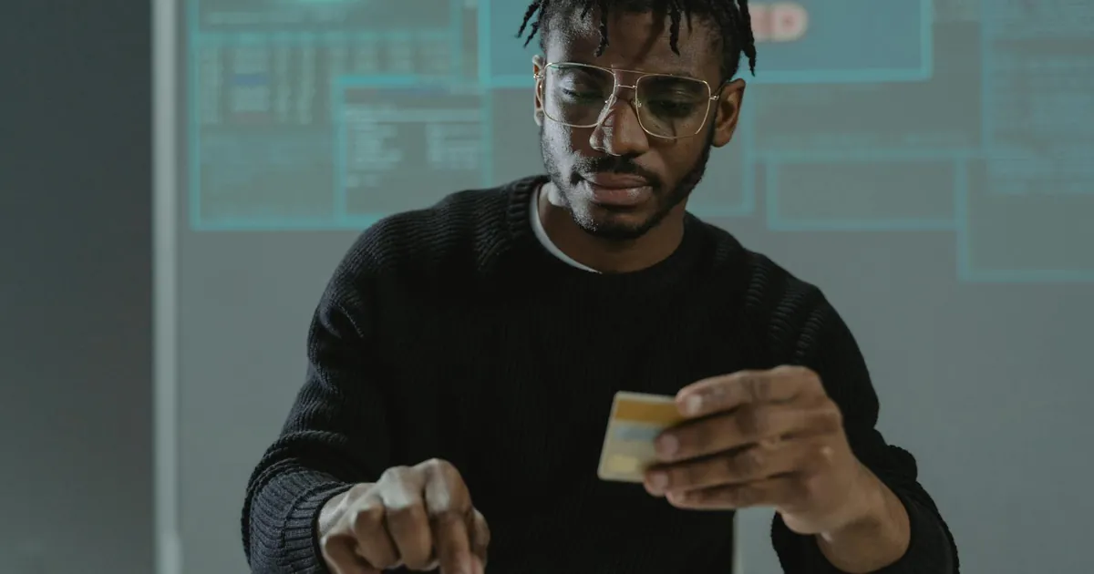 Man with glasses holding a bank card while typing on a keyboard, sitting in front of a computer.