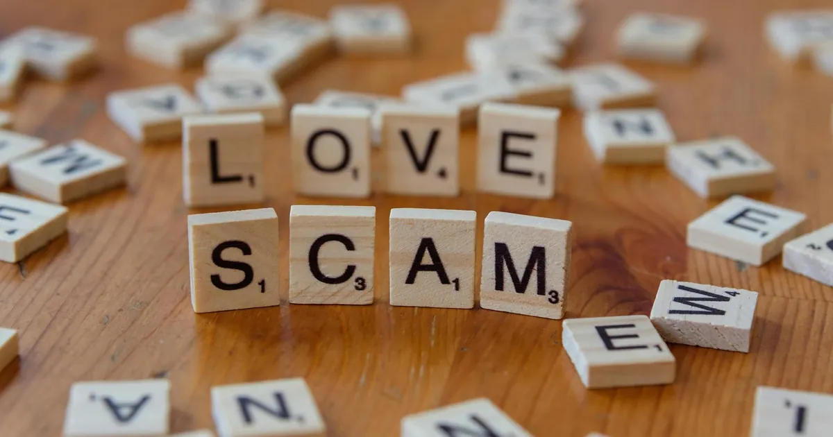 Scrabble tiles form words 'LOVE' and 'SCAM' on a wooden surface, illustrating deceit in relationships.