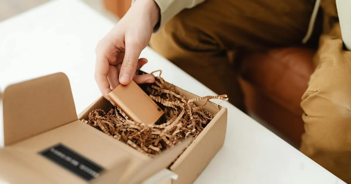 A person unboxes a small item from a cardboard package filled with crinkle paper, indoors.