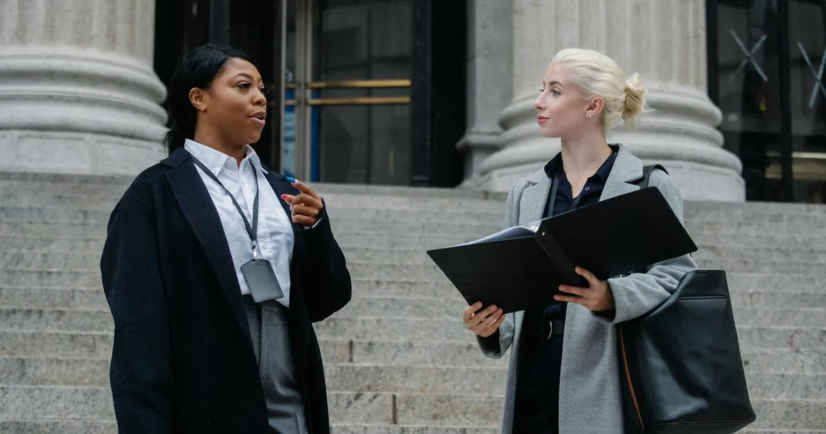 Two professional women in discussion on outdoor courthouse steps, holding documents.