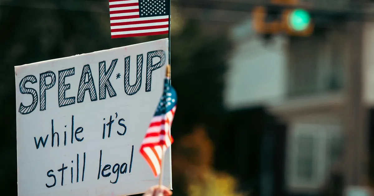 Protest sign with the message and American flag at a rally, highlighting free speech themes.