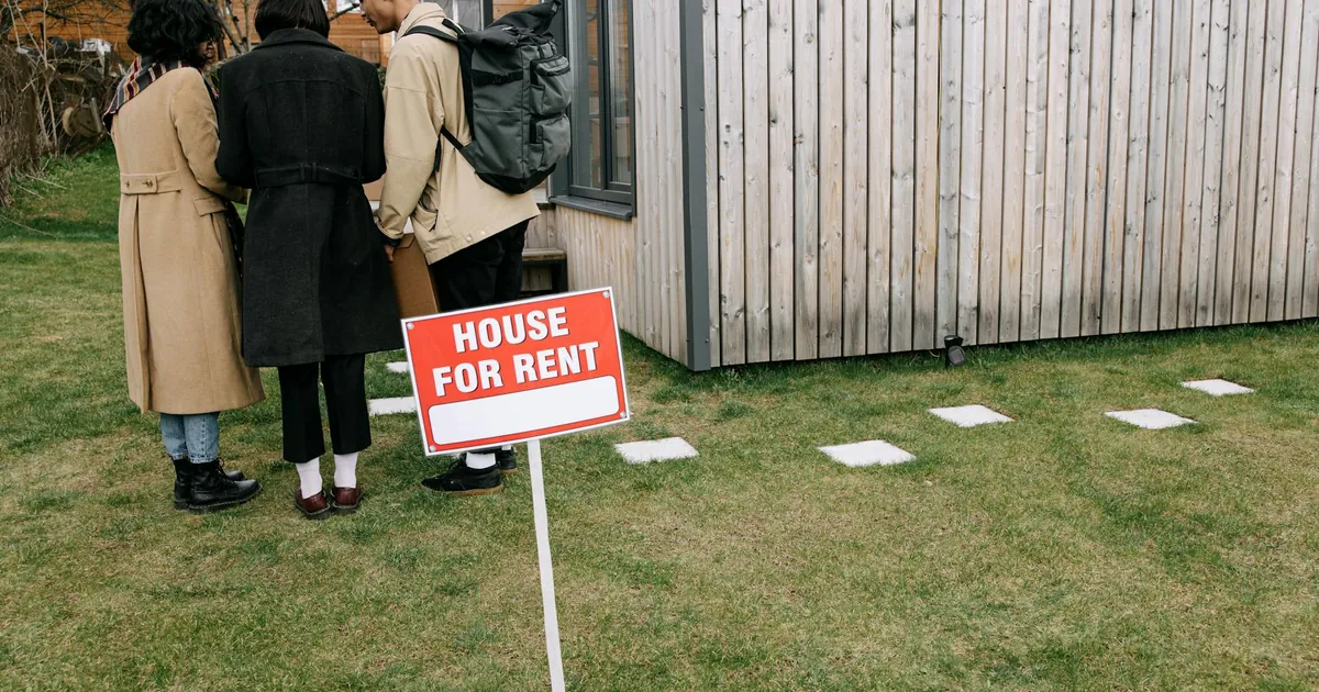 Group of adults viewing a wooden house with a 'House for Rent' sign on the lawn.