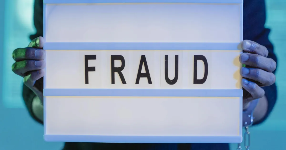 Close-up of hands holding a sign with 'fraud', illuminated in blue light. Close-up of hands holding a sign with 'fraud', illuminated in blue light.