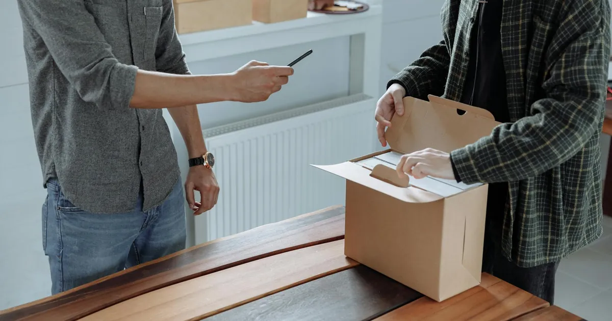 Two adults unpack and photograph a cardboard box using a smartphone indoors.