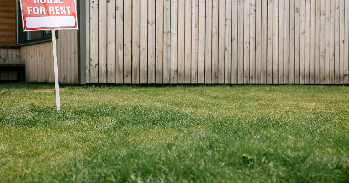 Close-up of 'House for Rent' sign on green lawn in front of a wooden house exterior.