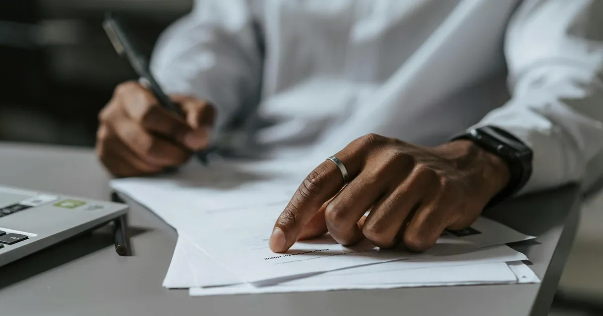 Close-up of a professional writing on documents at a work desk with a laptop.