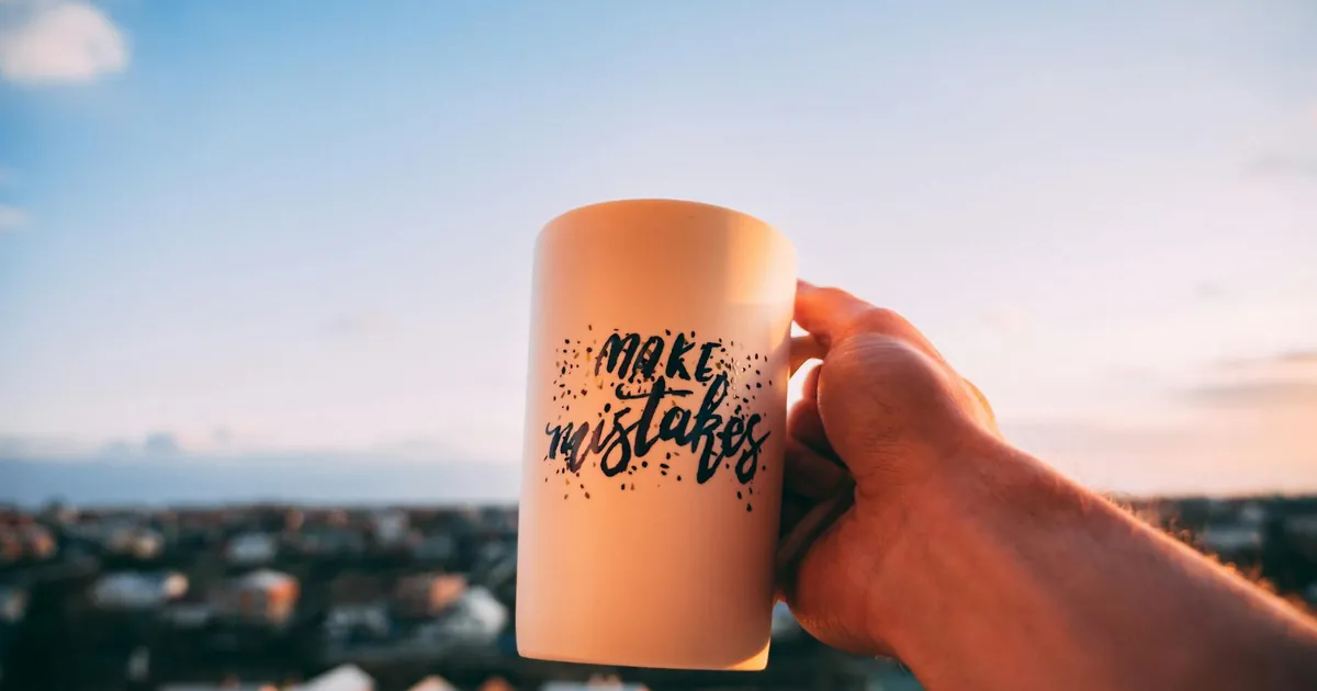 A hand holding a motivational mug with 'Make Mistakes' against a sunset in Lviv.